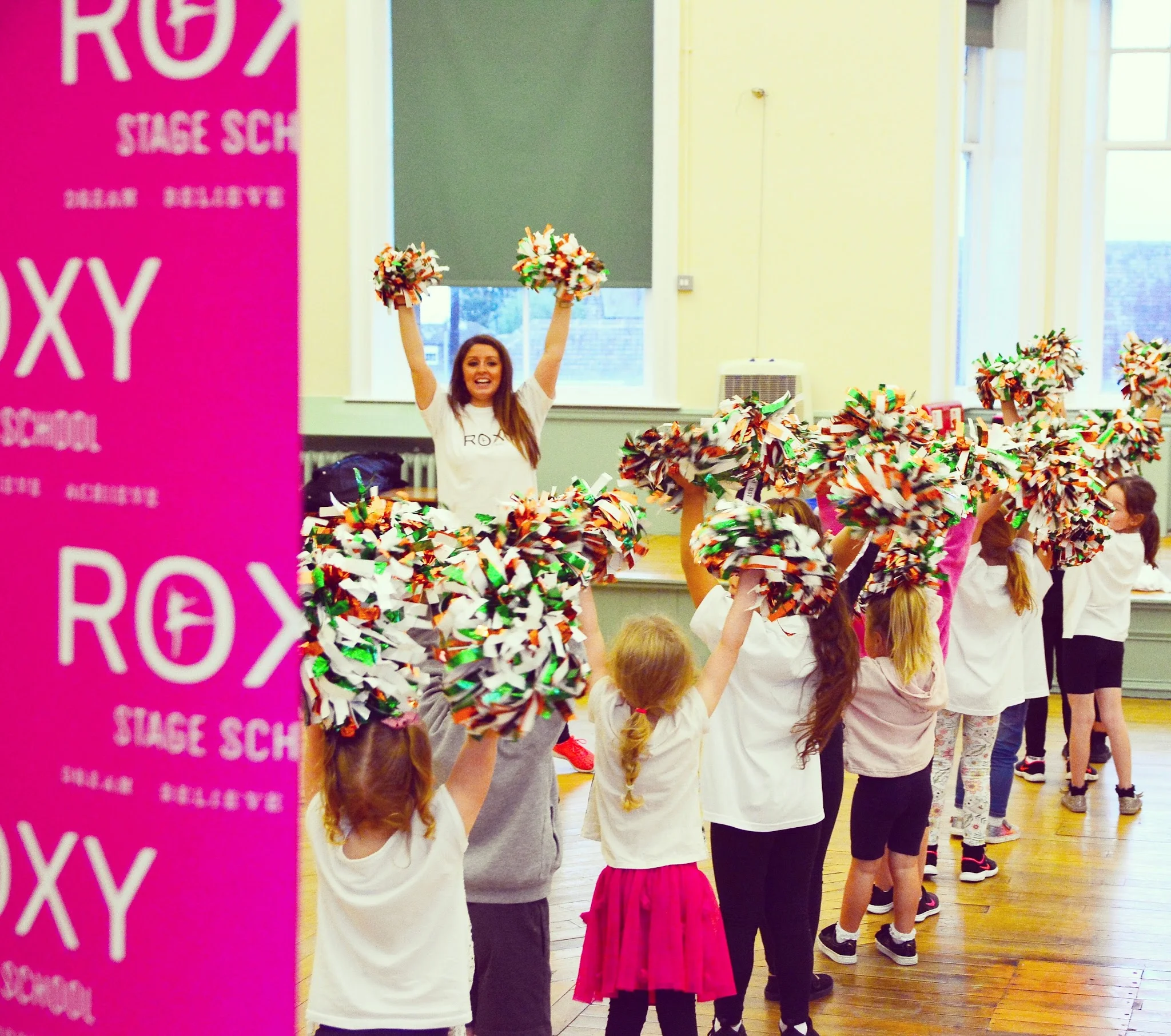 A Roxy Stage School class performing together with pom-poms beside the school's banners
